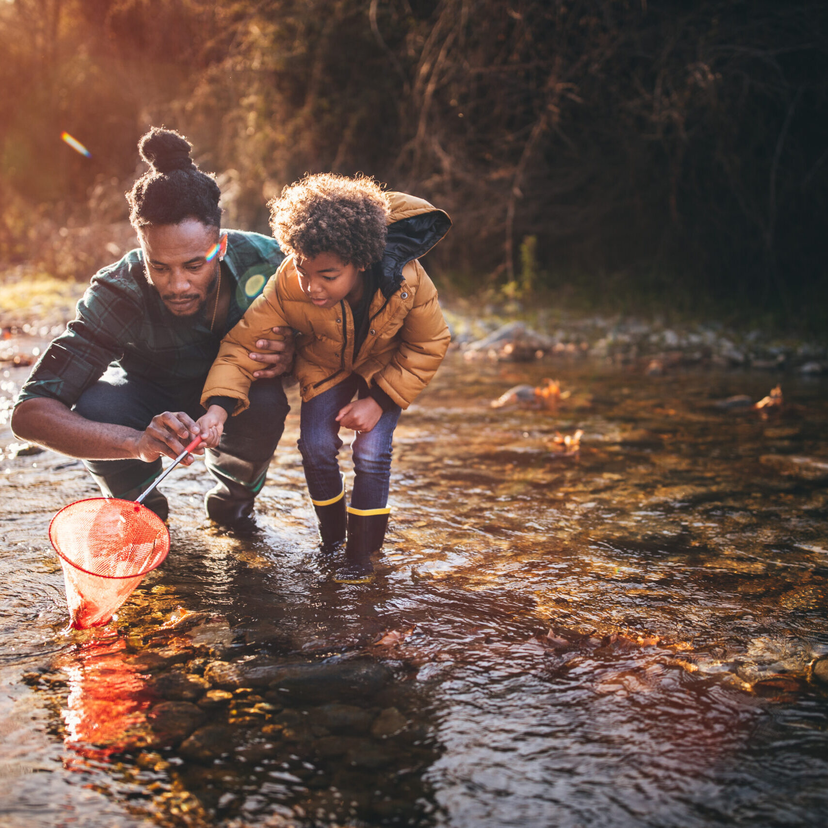 Young dad teaching son how to fish with fishing net in mountain stream at sunset